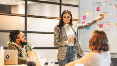 En réunion, une femme pointe du doigt un tableau blanc sur lequel des post-it sont classés par catégories